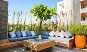 Elegante decoracion terrazas aticos con sofás de madera, cojines grises y un centro de mesa con fuego. Hierbas altas y un árbol crean un oasis urbano.
