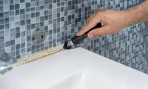 Mano usando una herramienta para quitar silicona vieja y sucia. El vinagre blanco puede ayudar a ablandar la silicona para facilitar su remoción.