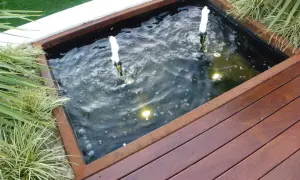 Un estanque jardín casero con dos surtidores de agua burbujeante, rodeado de vegetación y una tarima de madera.