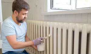Hombre con guantes pinta radiadores de hierro con brocha y pintura blanca.