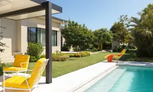 Un moderno jardín con piscina y porche, donde dos sillas amarillas invitan a relajarse bajo el sol.