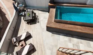 Terraza con tumbonas, mesa y barbacoa junto a una piscina elevada de obra con agua azul.