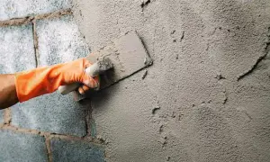 Mano con guante naranja aplica cemento para enfoscar pared exterior con llana.