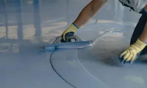 Persona con guantes amarillos aplica una capa de pintura azul sobre un suelo de terrazo con una llana.