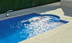 Un moderno patio con piscina de agua azul brillante, rodeado de baldosas de piedra y un seto verde. El sol crea destellos deslumbrantes en la superficie del agua.