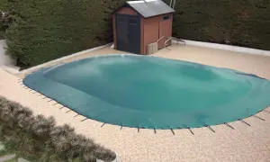 Cubierta verde protege la piscina para el invierno. Un cobertizo de madera y setos rodean el área.