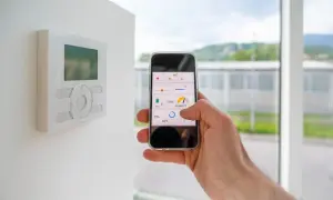 Control de domótica en casa con el móvil. Se ve un termostato en la pared y una mano manejando una app de control de clima y humedad.
