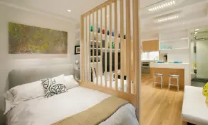 Un dormitorio moderno y funcional, parte de un diseño de interiorismo para estudios, con una cama confortable y una cocina integrada separada por una celosía de madera.