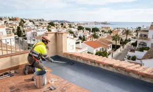 Trabajador aplica mortero bicapa en tejado con vistas a un pueblo costero.