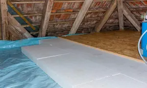 Aislamiento de suelo en una casa antigua con paneles de poliestireno sobre plástico azul. Vigas de madera y tejas visibles.
