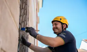 Hombre con casco amarillo sonríe mientras usa un pincel para rebozar paredes y rejas metálicas.