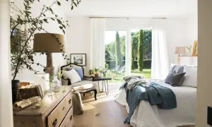 Acogedor dormitorio azul y blanco con cama, sillón y cómoda de madera. Ventanal con vistas a un jardín verde.