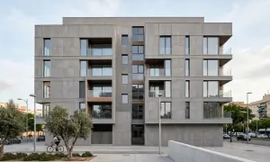 Edificio moderno con una elegante fachada ventilada de paneles grises, balcones acristalados y detalles en madera. Un ciclista pasa frente a árboles y la calle.