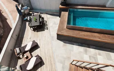 Terraza con tumbonas, mesa y barbacoa junto a una piscina elevada de obra con agua azul.