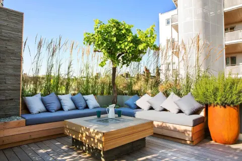 Elegante decoracion terrazas aticos con sofás de madera, cojines grises y un centro de mesa con fuego. Hierbas altas y un árbol crean un oasis urbano.