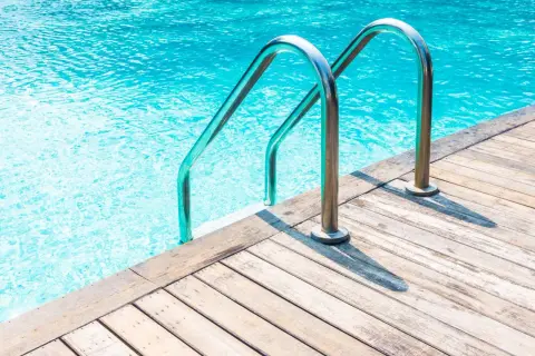 Escalera metálica para entrar a una alberca de agua cristalina, con borde de madera.