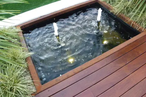 Un estanque jardín casero con dos surtidores de agua burbujeante, rodeado de vegetación y una tarima de madera.