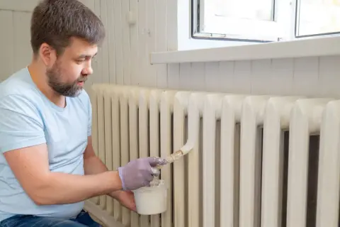 Hombre con guantes pinta radiadores de hierro con brocha y pintura blanca.