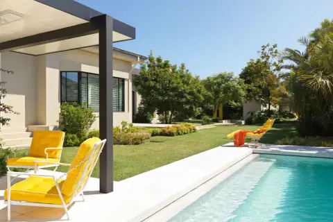 Un moderno jardín con piscina y porche, donde dos sillas amarillas invitan a relajarse bajo el sol.