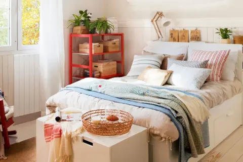 Acogedora habitación pequeña para adultos con cama, estantería roja y ventana luminosa.