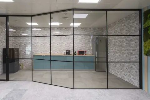 Una moderna cocina de oficina con una pared de cristal, gabinetes turquesas, electrodomésticos y una pared de ladrillos.
