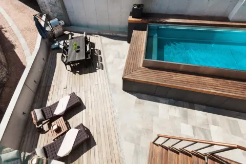 Terraza con tumbonas, mesa y barbacoa junto a una piscina elevada de obra con agua azul.