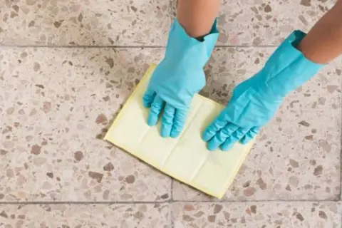Manos con guantes azules limpiando un suelo de terrazo con un paño amarillo.