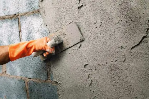 Mano con guante naranja aplica cemento para enfoscar pared exterior con llana.