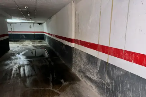 Pasillo de garaje con charcos en el suelo y marcas de humedad por filtración en las paredes pintadas con franjas rojas y grises.