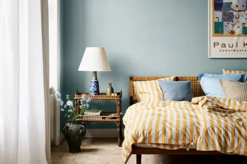 Habitación con paredes azul claro, cama con ropa de cama a rayas amarillas y blancas, y mesita de noche de ratán. Ideas de colores para habitaciones.