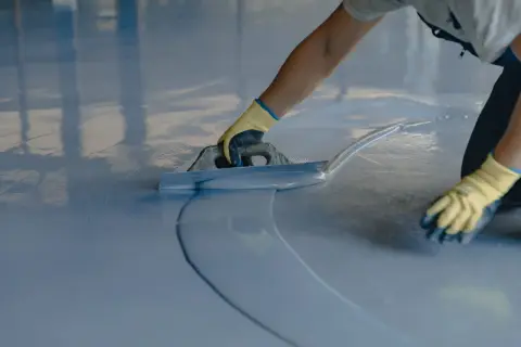 Persona con guantes amarillos aplica una capa de pintura azul sobre un suelo de terrazo con una llana.