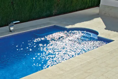 Un moderno patio con piscina de agua azul brillante, rodeado de baldosas de piedra y un seto verde. El sol crea destellos deslumbrantes en la superficie del agua.