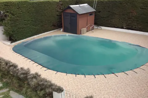 Cubierta verde protege la piscina para el invierno. Un cobertizo de madera y setos rodean el área.