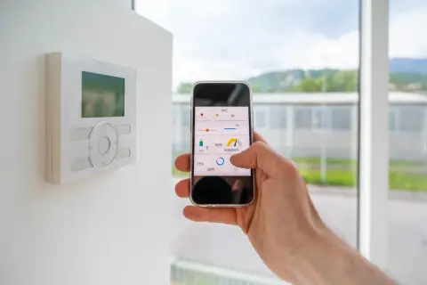 Control de domótica en casa con el móvil. Se ve un termostato en la pared y una mano manejando una app de control de clima y humedad.