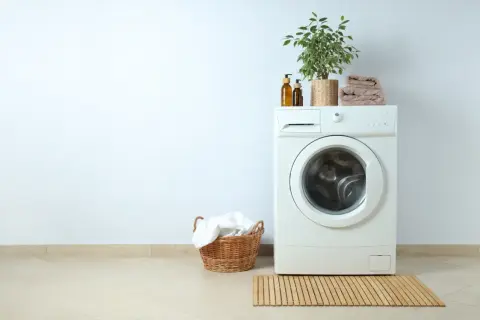 Lavadora blanca con planta y toallas encima. Cerca, una cesta con ropa blanca. La altura lavadora es perfecta para el espacio.