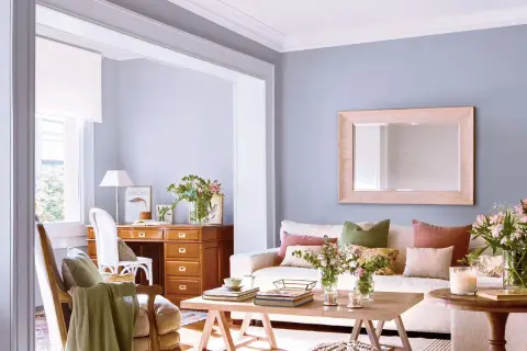 Ideas para decorar casa con poco dinero: sala acogedora con escritorio de madera, sofá blanco, cojines coloridos y flores frescas.