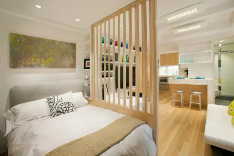 Un dormitorio moderno y funcional, parte de un diseño de interiorismo para estudios, con una cama confortable y una cocina integrada separada por una celosía de madera.