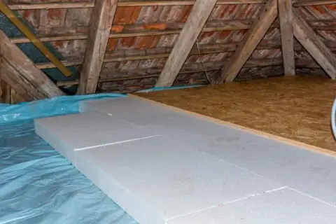 Aislamiento de suelo en una casa antigua con paneles de poliestireno sobre plástico azul. Vigas de madera y tejas visibles.