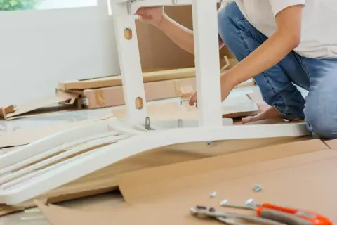 Persona ensambla mueble blanco. Se ve una pletina metálica uniendo piezas. Cajas y herramientas en el suelo.