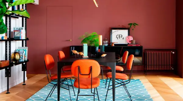 Comedor con paredes color terracota, sillas naranjas vibrantes y una alfombra azul con diseño de escamas. ¡Ideas de colores para pintar un salón audaz!