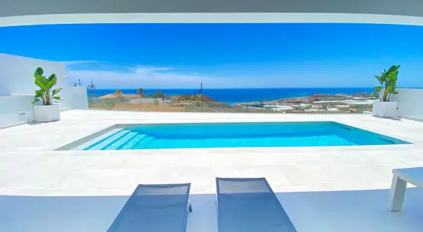 Dos tumbonas invitan a relajarse junto a una moderna piscina blanca, con el mar azul y el paisaje mediterráneo de fondo.