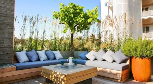 Elegante decoracion terrazas aticos con sofás de madera, cojines grises y un centro de mesa con fuego. Hierbas altas y un árbol crean un oasis urbano.