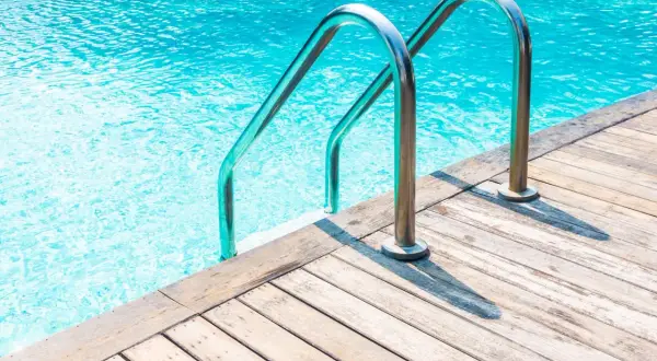 Escalera metálica para entrar a una alberca de agua cristalina, con borde de madera.