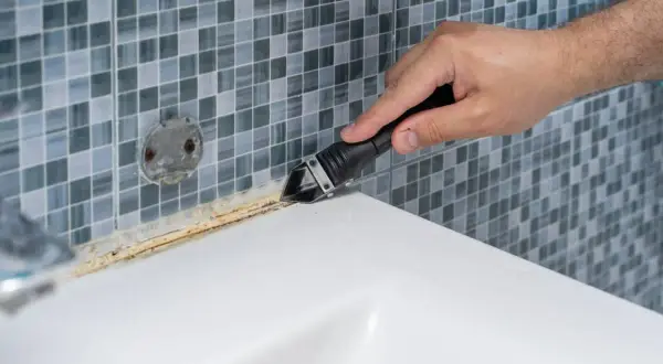 Mano usando una herramienta para quitar silicona vieja y sucia. El vinagre blanco puede ayudar a ablandar la silicona para facilitar su remoción.