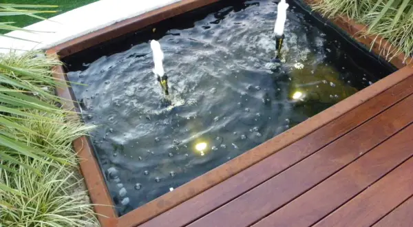 Un estanque jardín casero con dos surtidores de agua burbujeante, rodeado de vegetación y una tarima de madera.
