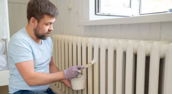 Hombre con guantes pinta radiadores de hierro con brocha y pintura blanca.
