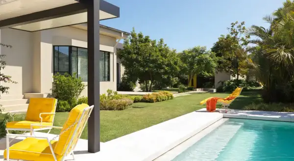 Un moderno jardín con piscina y porche, donde dos sillas amarillas invitan a relajarse bajo el sol.