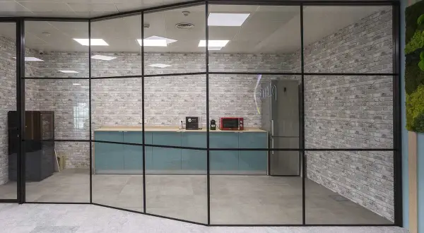 Una moderna cocina de oficina con una pared de cristal, gabinetes turquesas, electrodomésticos y una pared de ladrillos.