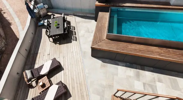 Terraza con tumbonas, mesa y barbacoa junto a una piscina elevada de obra con agua azul.
