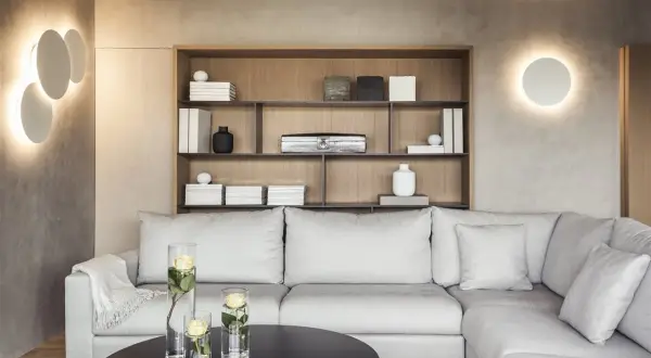 Iluminar un salón moderno con luces de pared circulares y una estantería de madera con objetos decorativos. Sofá gris y mesa negra con jarrones de rosas.